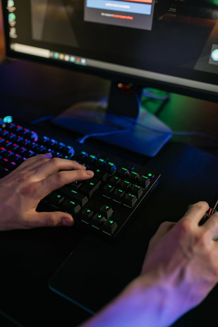 about-us-02 Close-up of hands using a keyboard and mouse on a gaming setup with colorful lights.